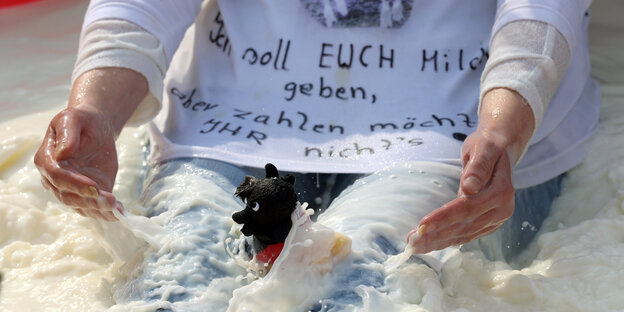 Ein Mensch in weißem T-Shirt und blauer Hose sitzt in Milch und hat einen kleinen schwarzen Spielzeughund im Schoß