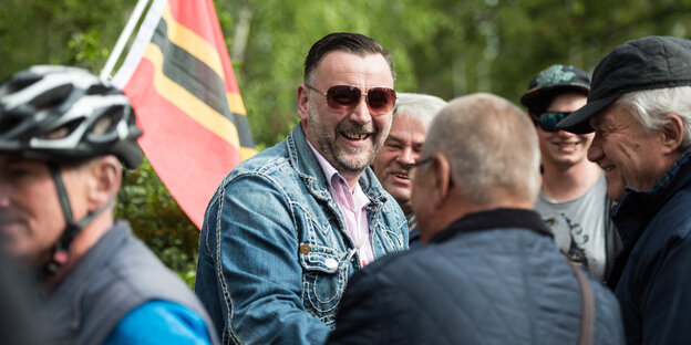 Mann mit Sonnenbrille, es ist Pegida-Gründer Lutz Bachmann, vor einer Fahne mit einem schwarz-rot-goldenen Kreuz, umringt von Anhängern Mann mit Sonnenbrille, es ist Pegida-Gründer Lutz Bachmann, vor einer Fahne mit einem schwarz-rot-goldenen Kreuz, umringt von Anhängern