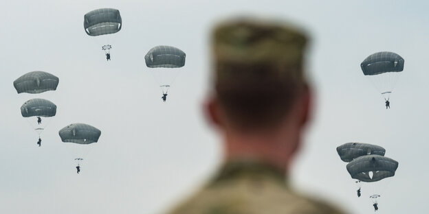 Ein Soldat blickt militärischen Fallschirmspringern entgegen, die im Landeanflug sind. Ein Soldat blickt militärischen Fallschirmspringern entgegen, die im Landeanflug sind.