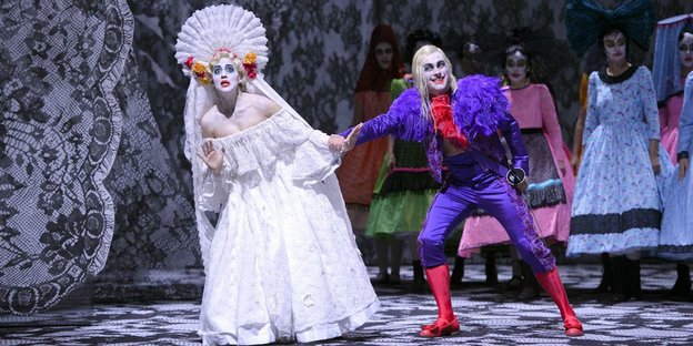 Eine Frau im weißen Kleid und ein Mann im lila Gewand auf einer Bühne