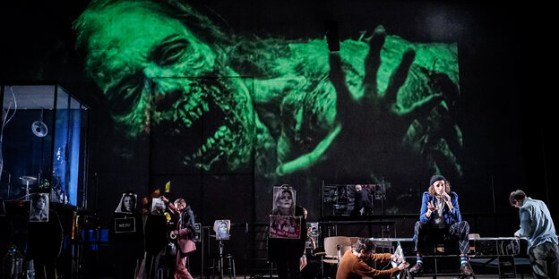 Eine Theaterbühne, auf der Menschen stehen, im Hintergrund eine grün-schwarze Projektion eines gruseligen menschlichen Monsters Eine Theaterbühne, auf der Menschen stehen, im Hintergrund eine grün-schwarze Projektion eines gruseligen menschlichen Monsters