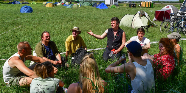 Ein Gruppe von Menschen sitzt auf einer Wiese, im Hintergrund sind Zelte zu sehen Ein Gruppe von Menschen sitzt auf einer Wiese, im Hintergrund sind Zelte zu sehen