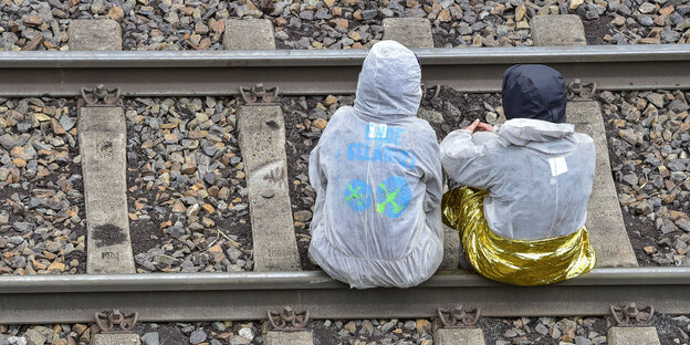 Zwei Menschen in weißen Overalls sitzen auf einem Bahngleis Zwei Menschen in weißen Overalls sitzen auf einem Bahngleis