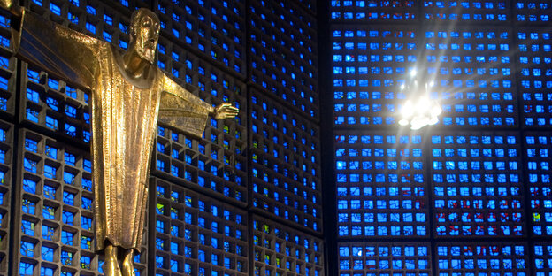 Eine goldene Jesus-Statue hängt vor einer blauen Glaswand. Eine goldene Jesus-Statue hängt vor einer blauen Glaswand.