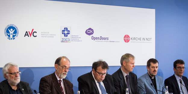 sechs Männer sitzen in einer Reihe, im Hintergrund verschiedene Logos christlicher Organisationen sechs Männer sitzen in einer Reihe, im Hintergrund verschiedene Logos christlicher Organisationen