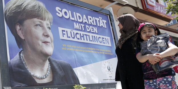 Auf einem Plakat ist Angela Merkel neben dem Satz „Solidarität mit Flüchtlingen“ zu sehen, eine Frau schaut auf das Plakat, daneben ein Kind Auf einem Plakat ist Angela Merkel neben dem Satz „Solidarität mit Flüchtlingen“ zu sehen, eine Frau schaut auf das Plakat, daneben ein Kind