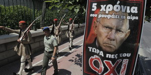 Das Bild Wolfgang Schäubles auf einem Plakat mit griechischer Schrift. Im Hintergrund marschieren Männer mit Waffe und in Uniform