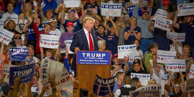 Ein Mann steht an einem Rednerpult und schreit ins Mikro. Es ist Donald Trump. Im Hintergrund sieht man seine Anhänger mit Schildern, auf denen sein Name steht Ein Mann steht an einem Rednerpult und schreit ins Mikro. Es ist Donald Trump. Im Hintergrund sieht man seine Anhänger mit Schildern, auf denen sein Name steht