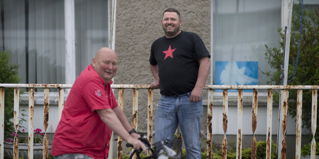 Zwei Männer, lachend vor einer Hausfassade. Einer der Männer sitzt auf einem Fahrrad Zwei Männer, lachend vor einer Hausfassade. Einer der Männer sitzt auf einem Fahrrad