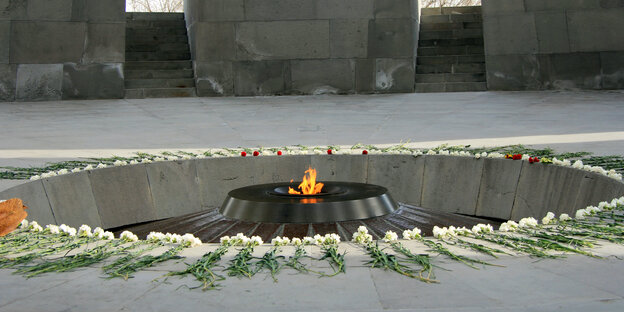 Foto aus einer Gedenkstätte: In der Mitte brennt ein Feuer, drum herum liegen kreisförmig viele Blumen Foto aus einer Gedenkstätte: In der Mitte brennt ein Feuer, drum herum liegen kreisförmig viele Blumen