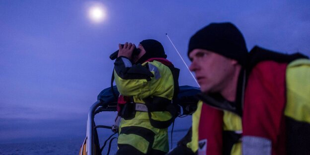 Zwei Männer mit Schwimmwesten in einem Boot auf dem Meer, einer schaut durch ein Fernglas Zwei Männer mit Schwimmwesten in einem Boot auf dem Meer, einer schaut durch ein Fernglas