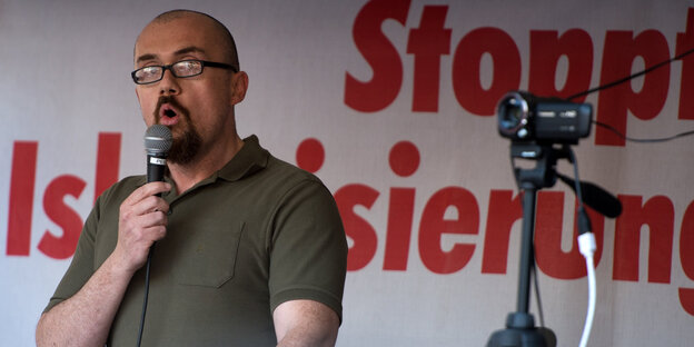 Ein Mann mit kurzen Haaren und Bart spricht in ein Mikrofon, im Hintergrund steht "Stoppt Islamisierung" Ein Mann mit kurzen Haaren und Bart spricht in ein Mikrofon, im Hintergrund steht "Stoppt Islamisierung"
