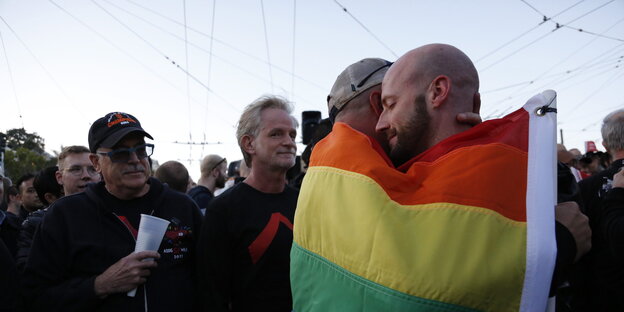 Zwei Männer umarmen sich. Sie haben eine Regenbogenfahne um sich gewickelt