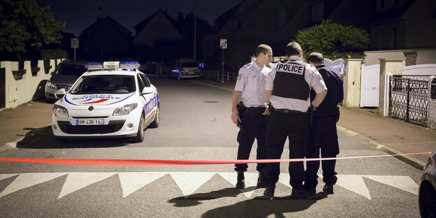 Drei Polizisten stehen neben einem Polizeiauto auf der abgesperrten Straße in Magnanville Drei Polizisten stehen neben einem Polizeiauto auf der abgesperrten Straße in Magnanville