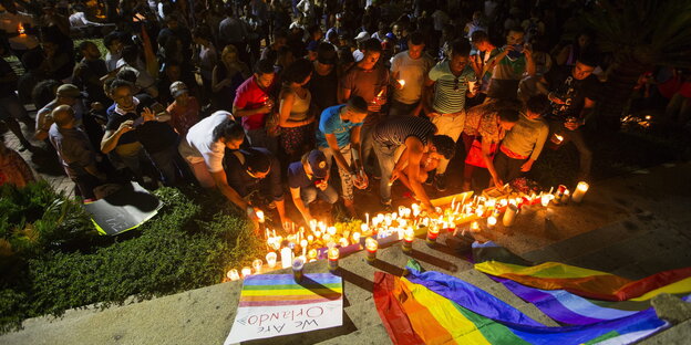 Viele Menschen zünden Kerzen an und stehen vor einer Regenbogen-Fahne Viele Menschen zünden Kerzen an und stehen vor einer Regenbogen-Fahne