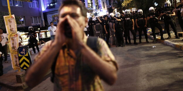 Im Hintergrund eine Polizeikette, im Vordergrund ein einzelner Demonstrant, der die Hände um den Mund legt und etwas ruft Im Hintergrund eine Polizeikette, im Vordergrund ein einzelner Demonstrant, der die Hände um den Mund legt und etwas ruft