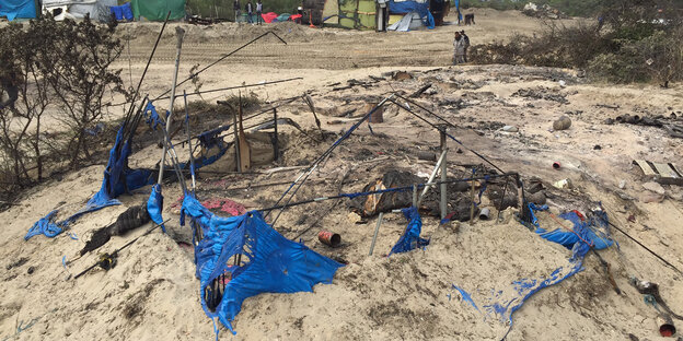 Auf einem Sandhügel liegen die Reste einer zerstörten Unterkunft: blaue Plastikfetzen und verkohlte Metallteile Auf einem Sandhügel liegen die Reste einer zerstörten Unterkunft: blaue Plastikfetzen und verkohlte Metallteile