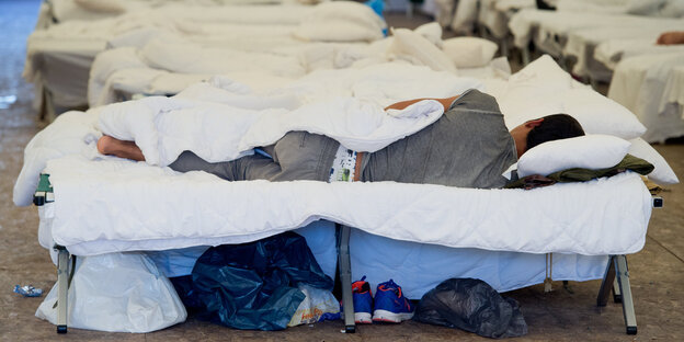 Ein Mensch liegt in einem Feldbett, um ihn herum stehen weitere Reihen von Feldbetten