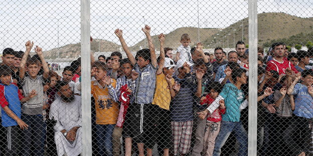 Menschen, darunter viele Kinder, hinter einem Zaun, im Hintergrund Hügel Menschen, darunter viele Kinder, hinter einem Zaun, im Hintergrund Hügel