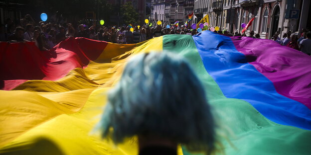 Eine Person mit türkisfarbenen Haar steht vor einer großen Regenbogenfahne Eine Person mit türkisfarbenen Haar steht vor einer großen Regenbogenfahne