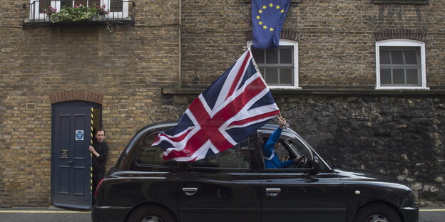 Aus einem britischen schwarzen Taxi hält jemand eine große britische Flagge. Über dem Auto hängt aus einem Fenster eine EU-Flagge Aus einem britischen schwarzen Taxi hält jemand eine große britische Flagge. Über dem Auto hängt aus einem Fenster eine EU-Flagge