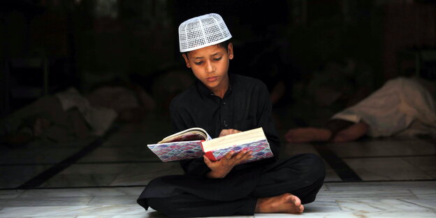 Ein Junge in dunklem Gewand und weißer Mütze ließt, mit überkreuzten Beinen sitzend, den Koran Ein Junge in dunklem Gewand und weißer Mütze ließt, mit überkreuzten Beinen sitzend, den Koran