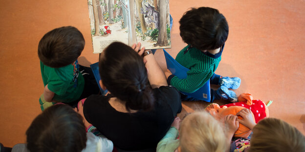 Eine Frau sitzt umringt von fünf Kinder und liest ihnen einen Buch vor – aus der Vogelperspektive betrachtet Eine Frau sitzt umringt von fünf Kinder und liest ihnen einen Buch vor – aus der Vogelperspektive betrachtet