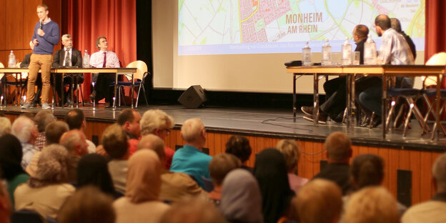 Eine Menschenmenge sitzt vor einer Bühne, auf der Menschen an Tischen sitzen und ein Mann vor den Tischen spricht Eine Menschenmenge sitzt vor einer Bühne, auf der Menschen an Tischen sitzen und ein Mann vor den Tischen spricht