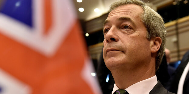 Links im Vordergrund die Flagge Großbritanniens, rechts Nigel Farage, ein älterer Mann mit grauen Haaren im Profil Links im Vordergrund die Flagge Großbritanniens, rechts Nigel Farage, ein älterer Mann mit grauen Haaren im Profil