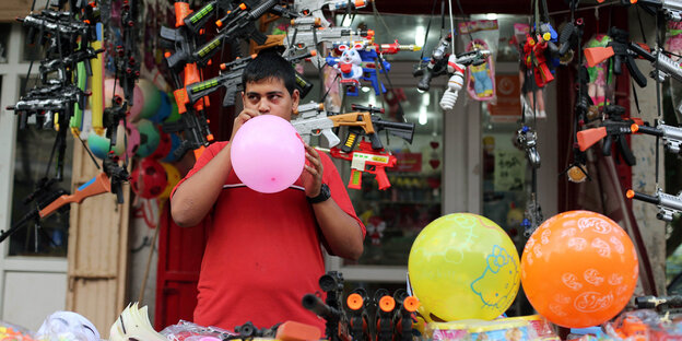 Ein Man bläst an einem Verkaufsstand Luftballons auf, hinter ihm hängen Spielzeugwaffen Ein Man bläst an einem Verkaufsstand Luftballons auf, hinter ihm hängen Spielzeugwaffen
