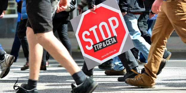 Menschen laufen über eine Straße. Eine Person hält ein Schild auf dem Stop TTIP und Ceta steht Menschen laufen über eine Straße. Eine Person hält ein Schild auf dem Stop TTIP und Ceta steht