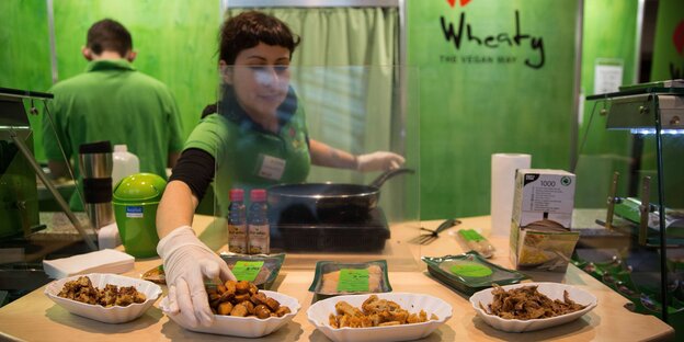 An einem grünen Messestand serviert eine Frau verschiedene Wurst-Proben An einem grünen Messestand serviert eine Frau verschiedene Wurst-Proben