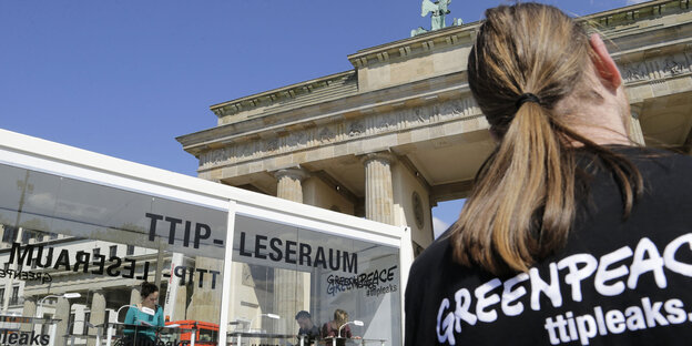 Szene vor dem Brandenburger Tor: Ein Glaskasten und ein Mensch von hinten mit langem Zopf, auf dem Tshirt die Aufschrift Greenpeace ttipleaks Szene vor dem Brandenburger Tor: Ein Glaskasten und ein Mensch von hinten mit langem Zopf, auf dem Tshirt die Aufschrift Greenpeace ttipleaks