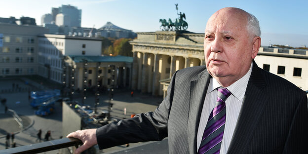 Ein älterer Mann mit Glatze in Anzug steht auf einem Balkon, im Hintergrund ist das Brandenburger Tor zu sehen Ein älterer Mann mit Glatze in Anzug steht auf einem Balkon, im Hintergrund ist das Brandenburger Tor zu sehen