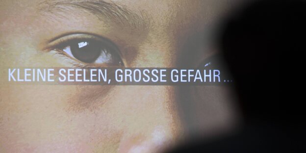 Kindersgesicht auf Leinwand mit Beschriftung „Kleine Seelen, große Gefahr“, davor Schatten des Kopfes eines erwachsenen Mannes Kindersgesicht auf Leinwand mit Beschriftung „Kleine Seelen, große Gefahr“, davor Schatten des Kopfes eines erwachsenen Mannes