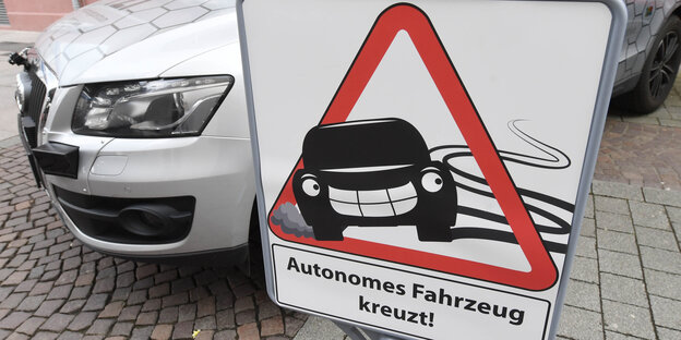 Auf einem Schild steht „Autonomes Auto kreuzt“. Dahinter steht ein Auto Auf einem Schild steht „Autonomes Auto kreuzt“. Dahinter steht ein Auto