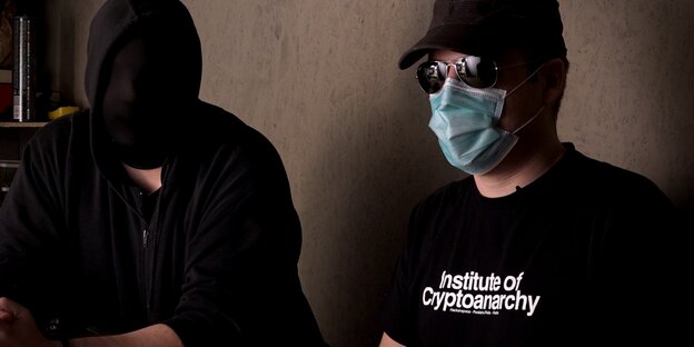 Zwei Menschen sitzen nebeneinander, einem hängt ein schwarzes Tuch über dem Kopf, der andere trägt Kappe, Sonnenbrille, Mundschutz und ein T-Shirt, auf dem „Institute of Cryptoanarchy“ steht Zwei Menschen sitzen nebeneinander, einem hängt ein schwarzes Tuch über dem Kopf, der andere trägt Kappe, Sonnenbrille, Mundschutz und ein T-Shirt, auf dem „Institute of Cryptoanarchy“ steht