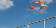 Ein Junge in Badehose, dessen Kopf vom Bildrand abgeschnitten ist, springt mit einem Ball in den Himmel