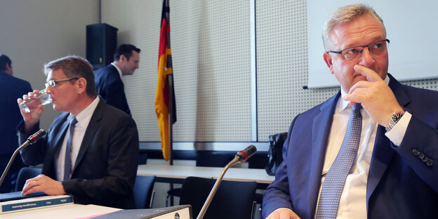 Zwei Männer sitzen an einem Konferenztisch, einer trinkt, der andere hat seine Hand im Gesicht Zwei Männer sitzen an einem Konferenztisch, einer trinkt, der andere hat seine Hand im Gesicht