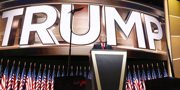 Donald Trump steht vor vielen Fahen und einem riesigen Schriftzug mit seinem Namen auf der Bühne in Cleveland Donald Trump steht vor vielen Fahen und einem riesigen Schriftzug mit seinem Namen auf der Bühne in Cleveland