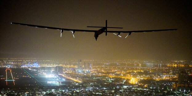 Ein Flugzeug fliegt über eine Stadt bei Nacht. Von unten leuchten die Lichter.