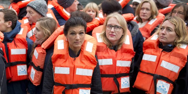 Eine Frau steht zwischen Menschen mit Rettungswesten. Es ist Sahra Wagenknecht. Ein Schild mit dem Schriftzug „Die Linke“ verdeckt ihren Mund Eine Frau steht zwischen Menschen mit Rettungswesten. Es ist Sahra Wagenknecht. Ein Schild mit dem Schriftzug „Die Linke“ verdeckt ihren Mund