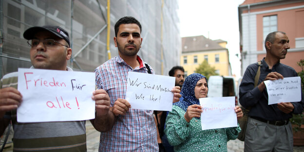 Menschen halten Zettel in die Höhe. Darauf steht unter anderem: „Frieden für alle“ und „Wir sind Menschen wie Ihr“