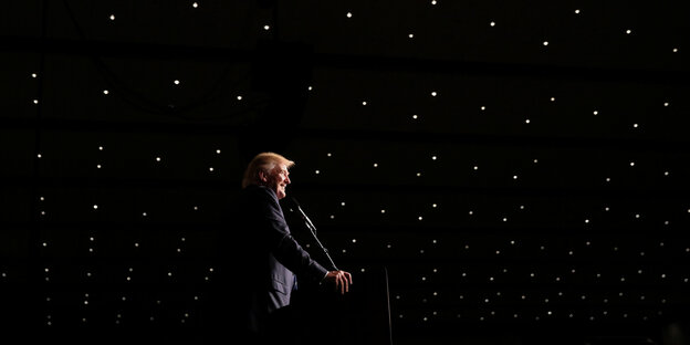 Ein Mann, es ist Donald Trump, steht am Rednerpult, um ihn herum ist es schwarz Ein Mann, es ist Donald Trump, steht am Rednerpult, um ihn herum ist es schwarz