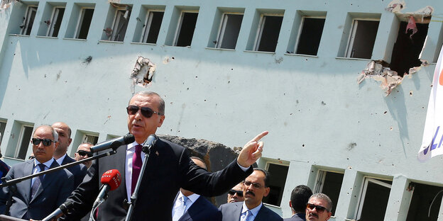 ein Mann in Anzug mit Sonnenbrille (es ist der türkische Präsident Erdogan) vor einem Haus mit Einschusslöchern ein Mann in Anzug mit Sonnenbrille (es ist der türkische Präsident Erdogan) vor einem Haus mit Einschusslöchern