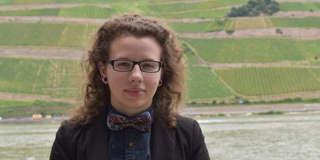 Eine junge Frau mit Locken und Brille steht vor einer grünen Hügellandschaft. Eine junge Frau mit Locken und Brille steht vor einer grünen Hügellandschaft.