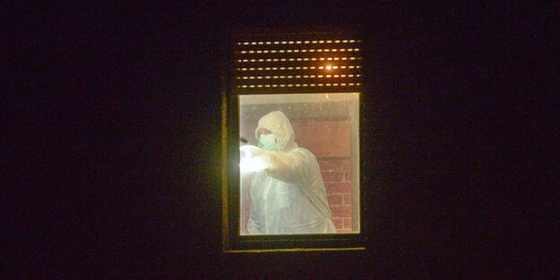 Das Bild zeigt ein Fenster während der Nacht. Im Lichtschein ist eine Person in einem weißen Ganzkörperanzug zu sehen Das Bild zeigt ein Fenster während der Nacht. Im Lichtschein ist eine Person in einem weißen Ganzkörperanzug zu sehen