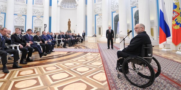 Craven spricht vor Publikum, im Hintergrund Putin Craven spricht vor Publikum, im Hintergrund Putin