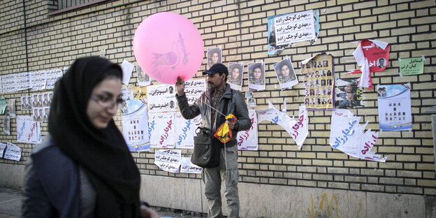 Ein Mann hält einen rosafarbenen Luftballon vor einer Wand, die mit Wahlpostern beklebt ist, im Vordergrund läuft eine Frau vorbei Ein Mann hält einen rosafarbenen Luftballon vor einer Wand, die mit Wahlpostern beklebt ist, im Vordergrund läuft eine Frau vorbei