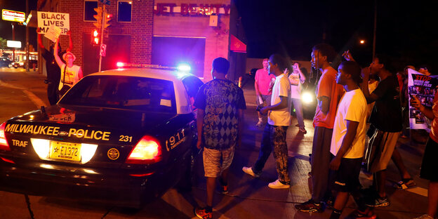 Es ist Nacht, links im Bild ein Polizeiauto und ein Mensch, der ein Schild hochhält, rechts neben dem Polizeiauto mehrere Menschen Es ist Nacht, links im Bild ein Polizeiauto und ein Mensch, der ein Schild hochhält, rechts neben dem Polizeiauto mehrere Menschen